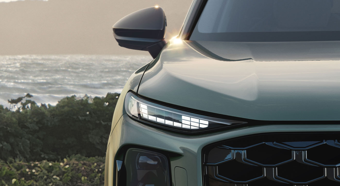Close-up of a sleek Audi Q3, green car front with a modern headlight design. The background shows a serene ocean view and greenery, creating a calm ambiance.